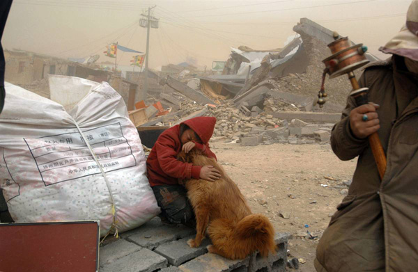 Ребенок обнимает свою собаку, сидя напротив разрушенного землетрясением дома